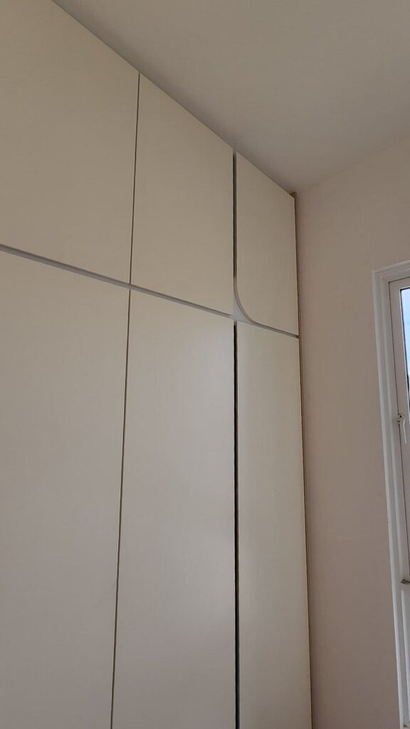 Full-height white built-in wardrobe with flush panels and rounded recessed handle detail, beside a tall window with greenery outside. Jade Hills, Kajang, Selangor.