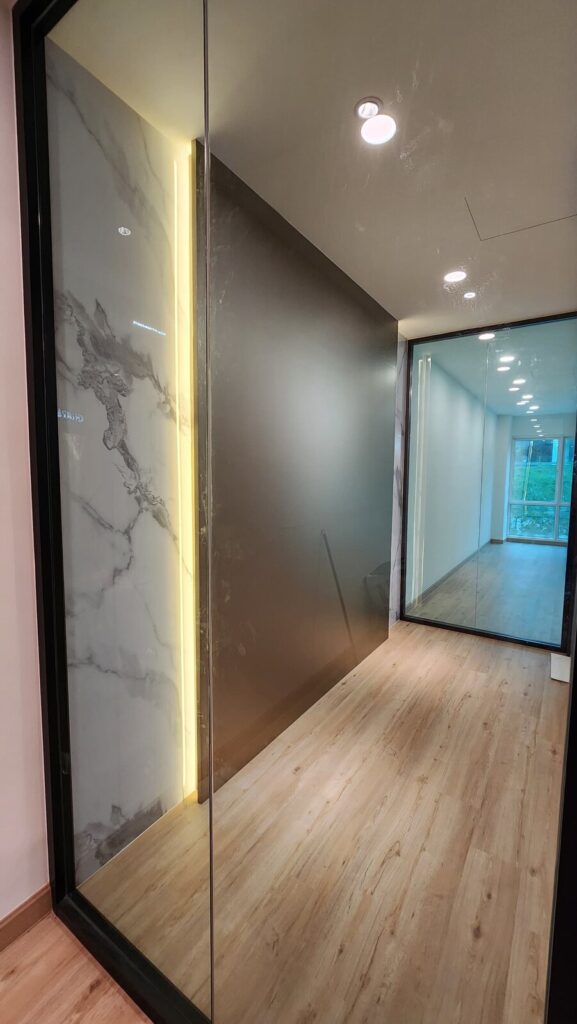 Hallway in a renovated office with marble-look feature wall, bronze frosted glass partition, glass-walled room with window and light wood flooring. Commercial Office Fit-Out, Kuala Lumpur.