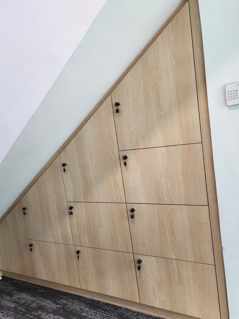 Completed triangular under-stair cabinet with locking drawers in light wood-grain laminate, installed against a pale wall beside a wall-mounted alarm keypad over carpeted flooring. Commercial Office Fit-Out, Kuala Lumpur.
