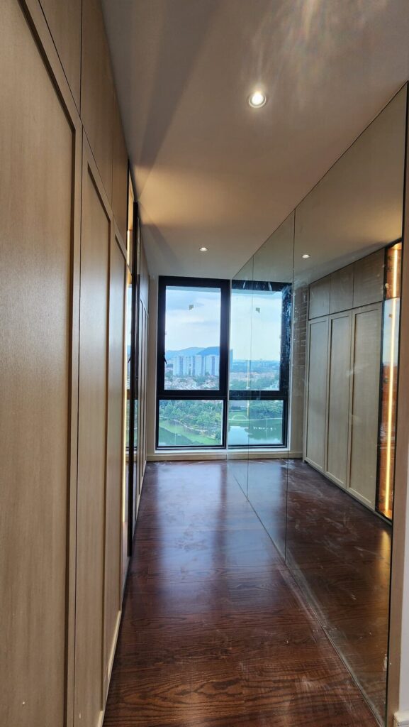 Walk-in wardrobe corridor with wood-veneer cabinetry, full-length mirror wall, dark wood floor and corner windows overlooking a lake and township. Park Regent, Desa ParkCity, Kuala Lumpur.