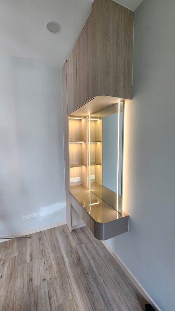 Built-in wood-grain dressing table unit with mirror, illuminated open cube shelving and a gold-toned countertop against pale green walls. Saujana Akasia, Shah Alam, Selangor.